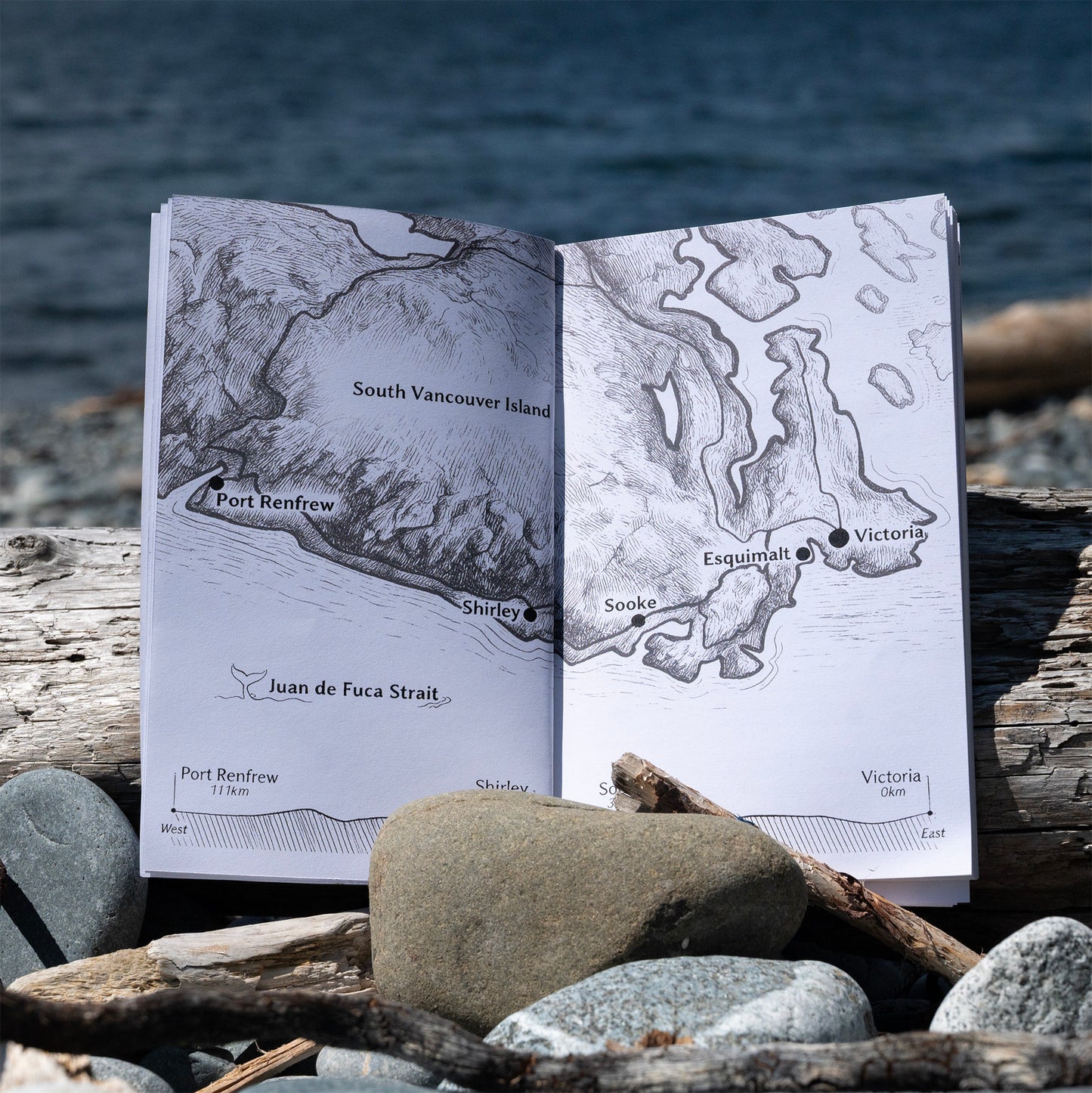 An open printed map booklet showing South Vancouver Island geography, placed on driftwood and rocks with the ocean in the background.