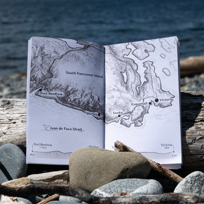 An open printed map booklet showing South Vancouver Island geography, placed on driftwood and rocks with the ocean in the background.