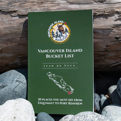 A green soft-covered book titled 'Vancouver Island Bucket List' by Juan de Fuca, standing upright outdoors among rocks in front of a weathered wooden wall. The cover features a circular Vancouver Island Bucket List logo and a white map outline.