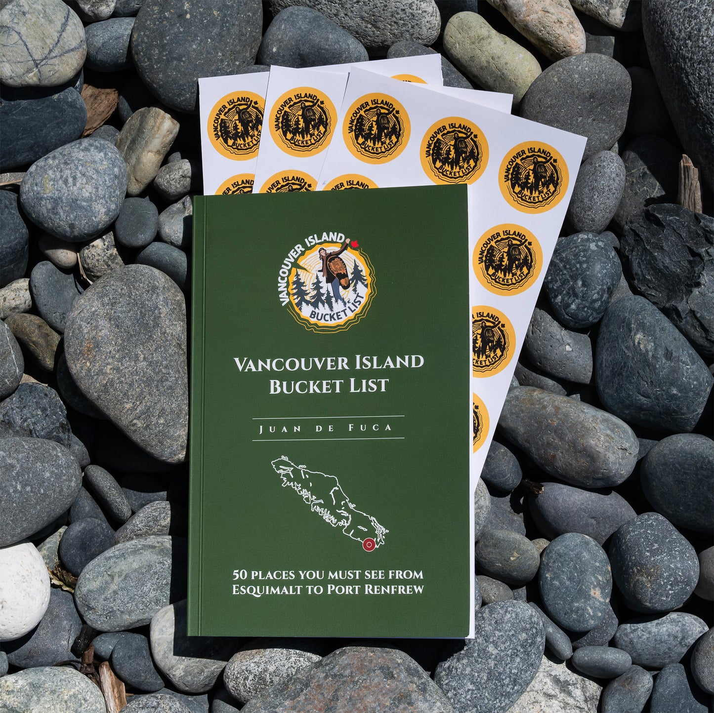 A green soft-cover book titled 'Vancouver Island Bucket List' by Juan de Fuca lies on a bed of dark gray river stones, with several printed sticker sheets fanned behind it.