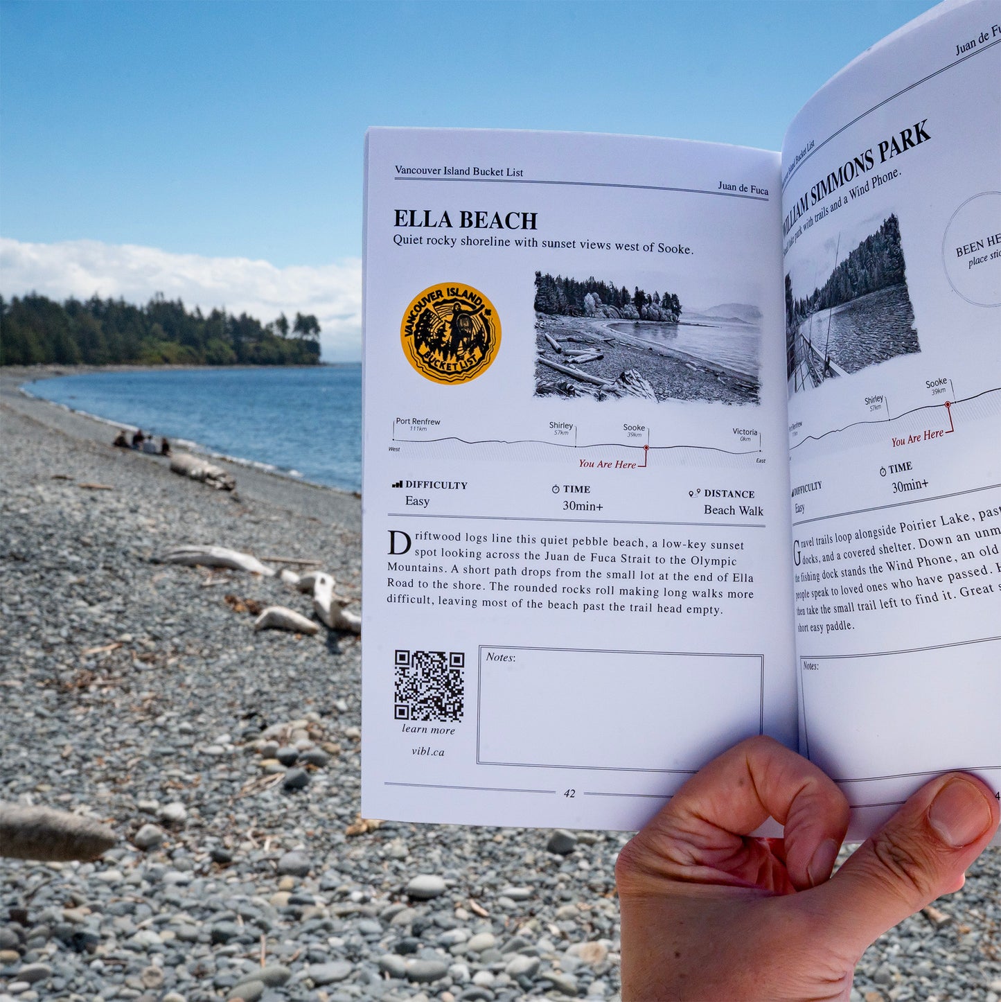 A hand holding open a printed booklet or guidebook about Ella Beach, with a rocky shoreline and blue water visible in the background. The booklet pages show maps, descriptive text, and a gold seal.