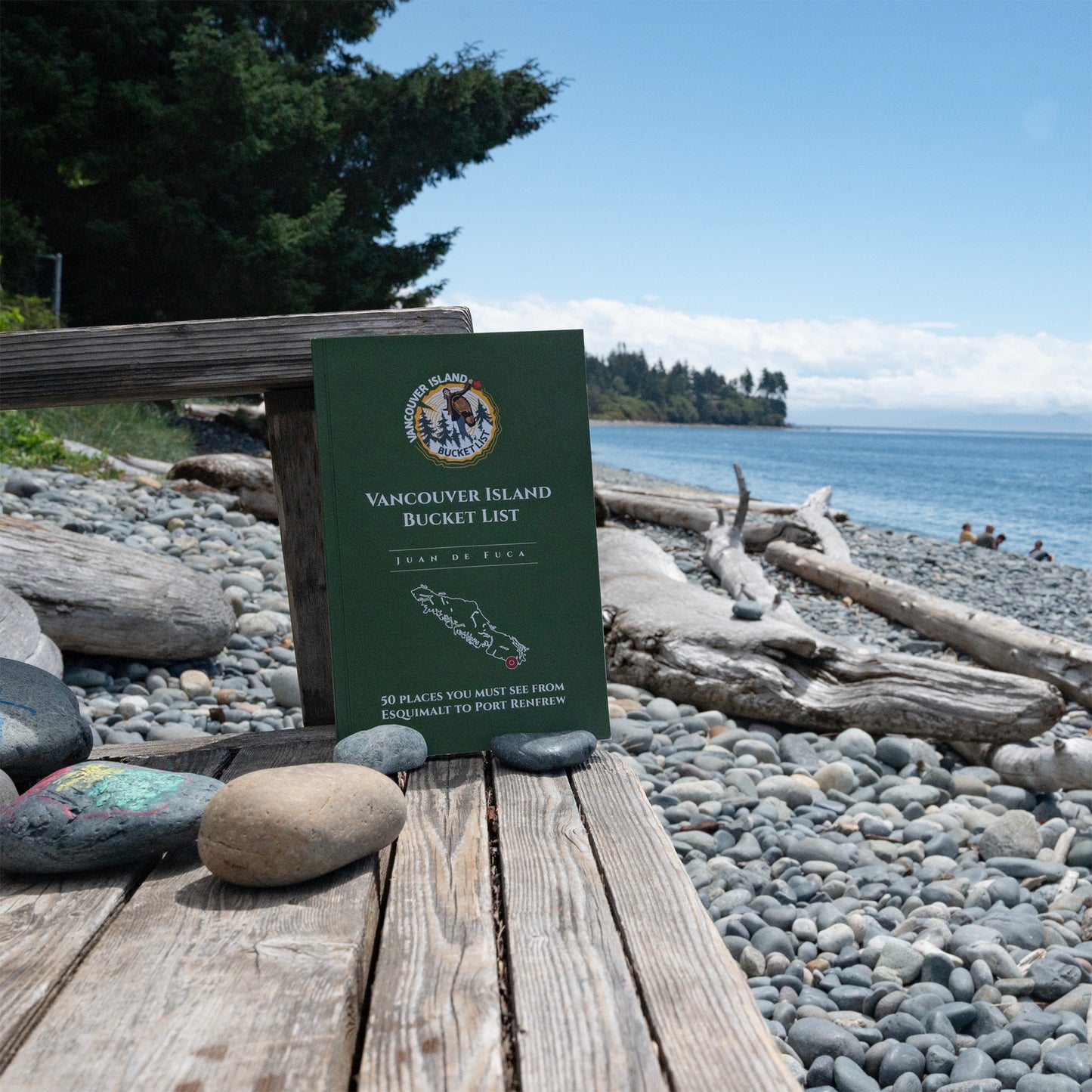 A green book titled 'Vancouver Island Bucket List' stands open on a wooden bench with a rocky beach and blue water in the background.